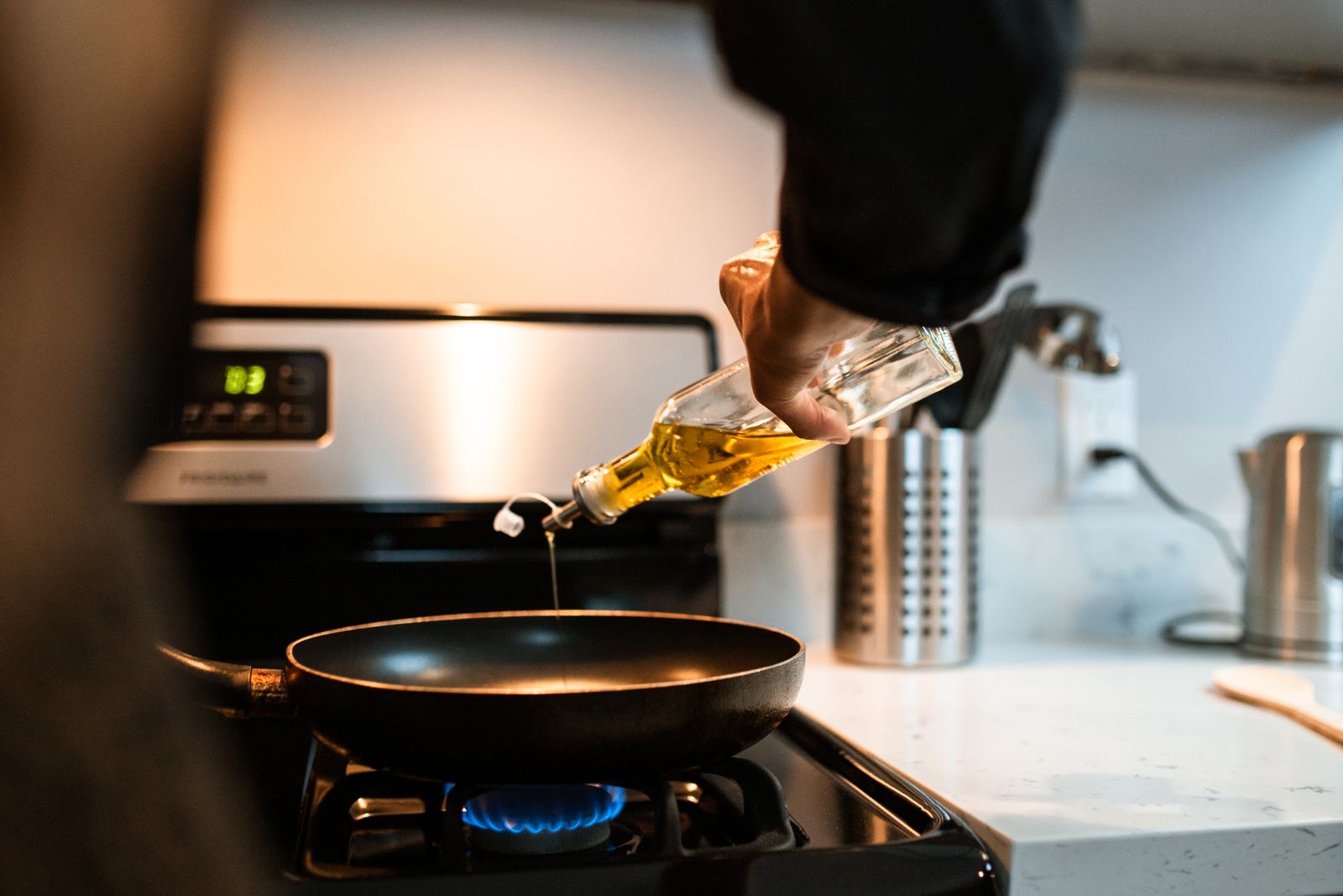 A hand pouring oil from a glass bottle into a frying pan on a gas hob